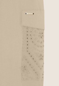 Tessuto beige con ricami floreali su una tasca; include un'etichetta metallica che legge "FREDDY." Texture liscia, design semplice.