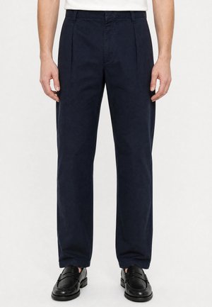 Homme portant un pantalon plissé bleu marine et des mocassins en cuir noir, debout devant un fond blanc.