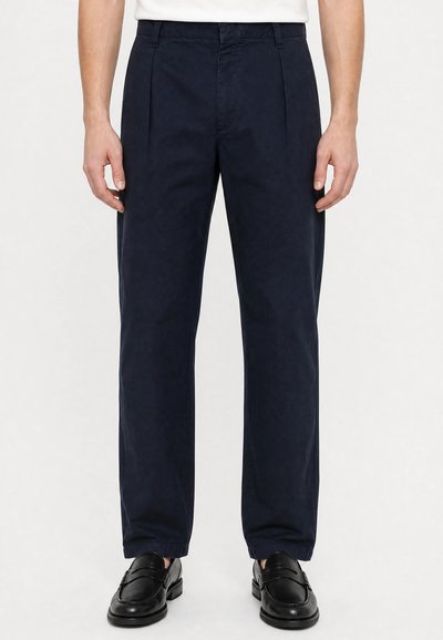 Homme portant un pantalon plissé bleu marine et des mocassins en cuir noir, debout devant un fond blanc.