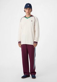 Witte trui met lange mouwen en bordeauxrode accenten, groene V-hals, gecombineerd met losvallende bordeauxrode broek met lichtblauwe strepen aan de zijkant en donkere schoenen.