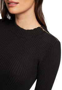 Haut en maille côtelée noire avec un col montant et un détail zippé sur l'épaule gauche. Le tissu a une texture lisse.