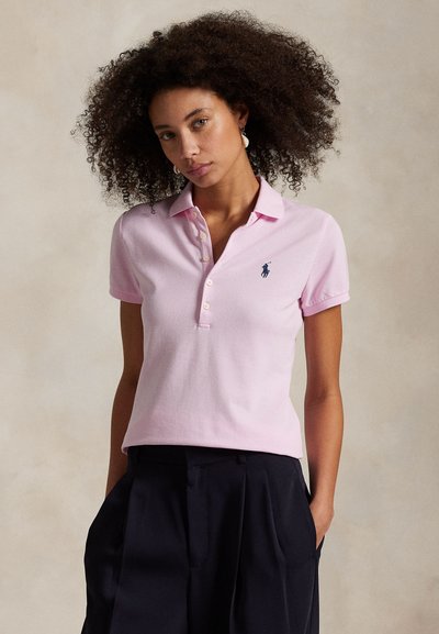 Woman with curly hair wearing a light pink short-sleeve polo shirt and black high-waisted pants, standing with hands in pockets.