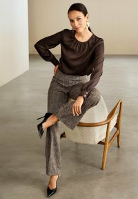 Frau in brauner Bluse und gemusterten Hosen, die auf einem beigen Armstuhl im mittleren Jahrhundert mit Holzrahmen sitzt und schwarze spitze Absätze trägt.