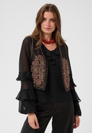 Femme portant un chemisier noir transparent avec des manches à volants et un gilet noir brodé, accessoirisée d’un collier rouge épais.