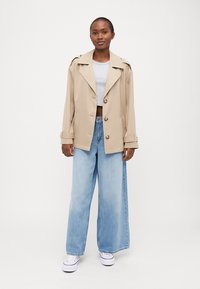 Beige dobbeltradet trenchcoat med store knapper, vide revers og oppbrettede ermer, kombinert med lyseblå vida bukser og hvite sneakers.