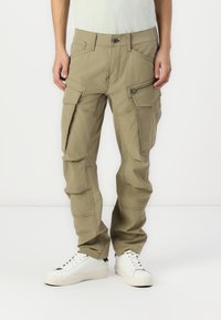 Beige cargo bukser med flere lommer, lynlåsdetaljer og forsterkede sømmer; brukt med hvite joggesko på en nøytral bakgrunn.