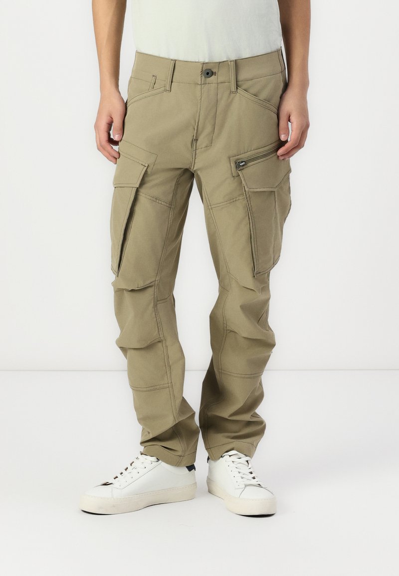 Beige cargo bukser med flere lommer, lynlåsdetaljer og forsterkede sømmer; brukt med hvite joggesko på en nøytral bakgrunn.