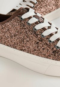 Prim-plan al unei adidași acoperiți cu sclipici maro, cu șireturi albe și talpă groasă din cauciuc alb, pe un fundal deschis.