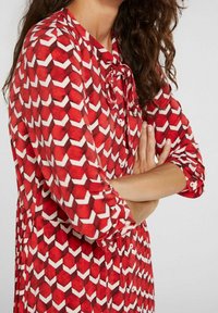 Blouse à motifs rouges avec un design géométrique et des accents blancs, fermeture à boutons, et un lien au niveau du col, fabriquée à partir de tissu léger.