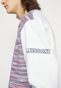 Maglione bianco con un design a strisce orizzontali multicolori lavorato a maglia sulle spalle. "MISSONI" stampato in blu navy sulla manica sinistra.