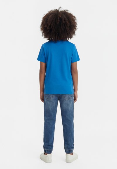 Παιδί με σγουρά μαλλιά που φορά μπλε μπλουζάκι, γυρισμένο τζιν και λευκά αθλητικά παπούτσια, στέκεται με την πλάτη προς τα έξω σε απλό φόντο.