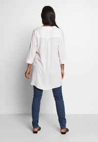 Túnica blanca de manga larga con dobladillo redondeado, que presenta un detalle de pliegue en la parte posterior, combinada con jeans skinny de color azul oscuro y sandalias negras.
