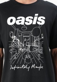 Sort bomulds t-shirt med fed hvid tekst "oasis" og en linjetegning af en stue med figurer og forskellige møbler.