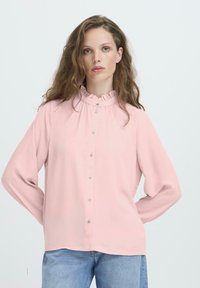 Vrouw met golvend bruin haar draagt een lichtroze blouse met knopen en een gerimpelde kraag, samen met lichtblauwe jeans, staande tegen een effen achtergrond.
