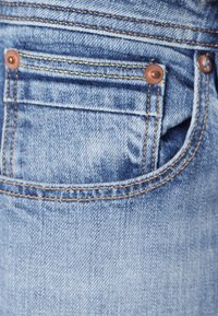 Jeans de mezclilla azul claro con costuras visibles, que cuentan con un bolsillo trasero con remaches de cobre y un diseño de bolsillo curvado.