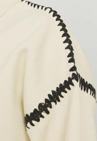 Krämfärgad tyg med texturerad yta, med en framträdande svart dekorativ söm längs axelsömmen.