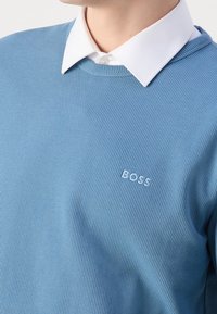 Hellblauer, strukturiert gestrickter Pullover mit einem weißen Hemd darunter. Kleines weißes "BOSS"-Logo auf der linken Brustseite.