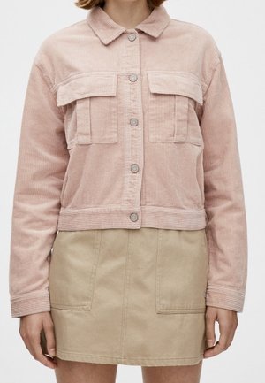 Femme portant une veste en velours côtelé rose clair avec des boutons et des poches à rabat, associée à une jupe beige avec des poches plaquées à l'avant.