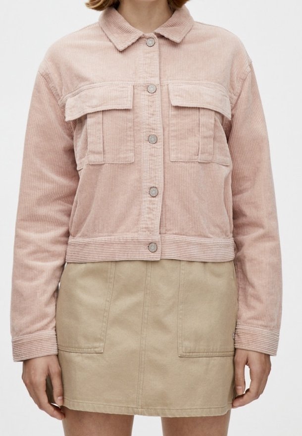 Femme portant une veste en velours côtelé rose clair avec des boutons et des poches à rabat, associée à une jupe beige avec des poches plaquées à l'avant.