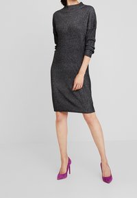 Abito a maglia aderente nero con scollo rotondo e maniche lunghe. Presenta una leggera lucentezza. Abbinato a scarpe con tacco alto in suede viola.