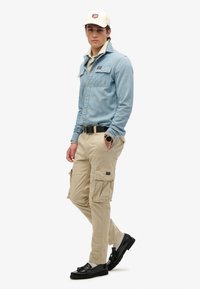 Lichtblauwe spijkerblouse met twee borstzakken, beige cargo broek met zijzakken, zwarte riem en zwarte schoenen. Crèmekleurige pet.