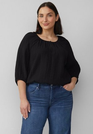 Schwarzes strukturiertes Oberteil mit rundem Halsausschnitt und Drei-Viertel-Ärmeln, kombiniert mit blauen Jeans. Wichtige Details sind die vertikalen Streifen auf dem Material.