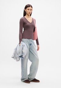 Top mauve cu mânecă lungă și decolteu în V, asortat cu blugi albi cu picioare largi de culoare albastru deschis. Ținând o jachetă din denim de culoare albastru deschis și purtând adidași maro.