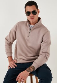 Sudadera de cremallera en beige claro de tejido suave, con un corte relajado y mangas largas. Modelo usando gafas de sol, sentado en un taburete de madera.