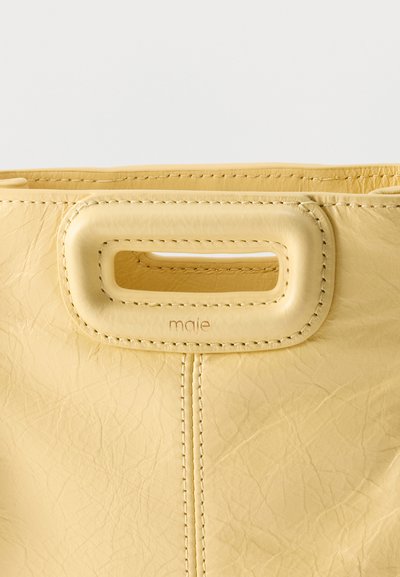 Κίτρινη δερμάτινη τσάντα χειρός με ορθογώνια λαβή με κοπή και ορατές ραφές, εμφανίζει ελαφριά υφή και το ανάγλυφο λογότυπο "maje" κάτω από τη λαβή.