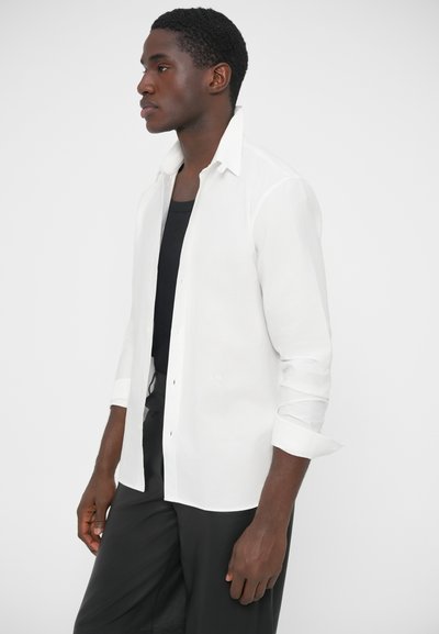 Chemise blanche à manches longues avec col boutonné, coupe slim, en tissu léger. Portée sur un débardeur noir, manches retroussées.