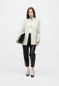 Γυναίκα που φοράει μπεζ σακάκι, μαύρο δερμάτινο παντελόνι και μαύρες γόβες, κρατώντας μια μεγάλη μαύρη τσάντα χειρός μπροστά σε απλό λευκό φόντο.