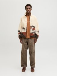 Jachetă din fleece crem cu un model montan în nuanțe de marron și gri, asortată cu un tricou marron și pantaloni lejeri din kaki, completați cu încălțăminte marron.