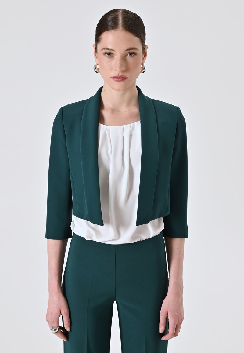 Giacca teal cropped con maniche a tre quarti, indossata sopra una blusa bianca plissettata. L'outfit è abbinato a pantaloni teal coordinati.