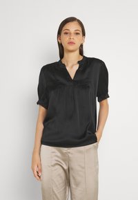 Black satin top with a ruffled neckline and short, gathered sleeves. The fabric has a smooth texture and a relaxed fit. Paired with beige pants.