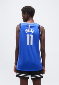 Άτομο που κοιτάζει προς τα πίσω, φορώντας μπλε φανέλα μπάσκετ με την επιγραφή "Irving 11" και μαύρο σορτς με λευκές ρίγες, πάνω σε απλό φόντο.