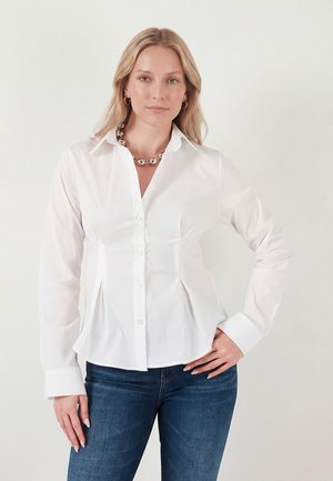 Blondína s rozopnutými vlasmi nosí priliehavú bielu košeľu na gombíky, tmavé džínsy, strieborný perlový náhrdelník a kruhové náušnice, pózuje s rukou na boku.