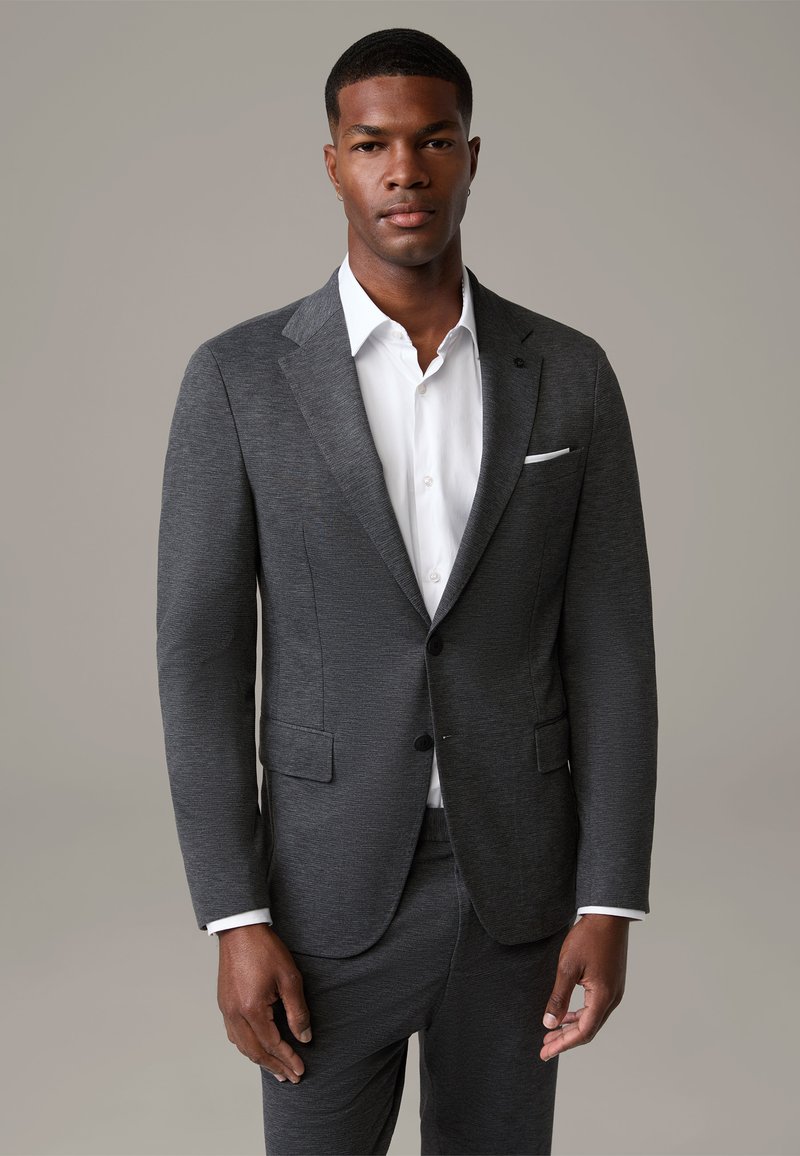 Charcoal grijs op maat gemaakt colbert met notch lapels, sluiting met één knoop, en zijzakken, gedragen over een witte overhemd met kraag.
