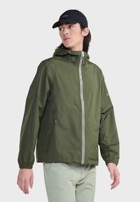 Oliivinvihreä vedenpitävä takki hupulla, joustavilla kalvosimilla ja valkoisella vetoketjulla. Päällä beigejen housujen kanssa ja yhdistetty tummansiniseen lippikseen.