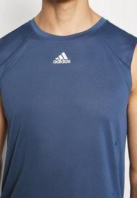 Sportovní tílko tmavě modré barvy, krátké rukávy, prodyšná látka s texturovaným vzorem, s bílým logem Adidas na hrudi.