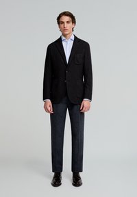 Blazer texturizado oscuro con un bolsillo de parche, combinado con pantalones oscuros y zapatos de cuero marrón. Camisa azul clara visible debajo de la chaqueta.