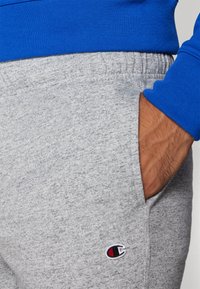 Gray sweatpants made of soft fabric with an elastic waistband and side pockets. Features a small embroidered logo in red and navy.