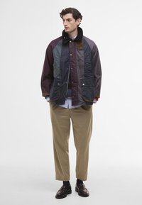 Többszínű dzseki, amely különböző anyagok összevonását mutatja, beleértve a fényes és matt textúrákat, műszőrme gallérral, két elülső zsebbel és laza szabással.