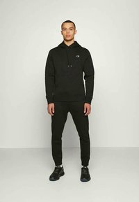 Μαύρη βαμβακερή φούτερ με μπροστινή τσέπη και κορδόνια, συνδυασμένη με μαύρες φόρμες. Το σύνολο ολοκληρώνεται με μαύρα αθλητικά παπούτσια.