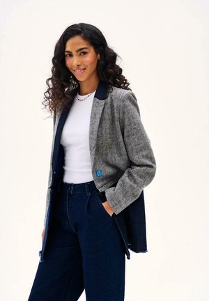 Kobieta o kręconych włosach ubrana w biały top, szarą marynarkę z niebieskim guzikiem i ciemnoniebieskie spodnie, uśmiechająca się z jedną ręką w kieszeni.