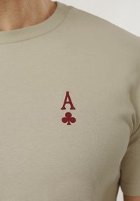 Μπεζ μπλουζάκι με στρογγυλή λαιμόκοψη, με ένα μικρό κόκκινο σύμβολο ασοβάτου μπαστούνι εκτυπωμένο στο αριστερό μέρος του στήθους.