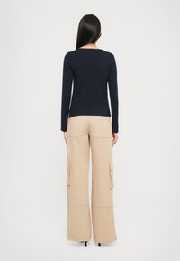 Femeie cu păr lung și închis la culoare, îmbrăcată cu un top lung cu mânecă navy și pantaloni cargo bej cu croială largă, stând cu fața la un fundal alb simplu.