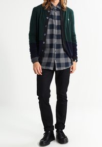 Groene en marineblauwe cardigan met knopen, over een grijs geruit shirt. Zwarte broek en zwarte schoenen maken de outfit compleet.