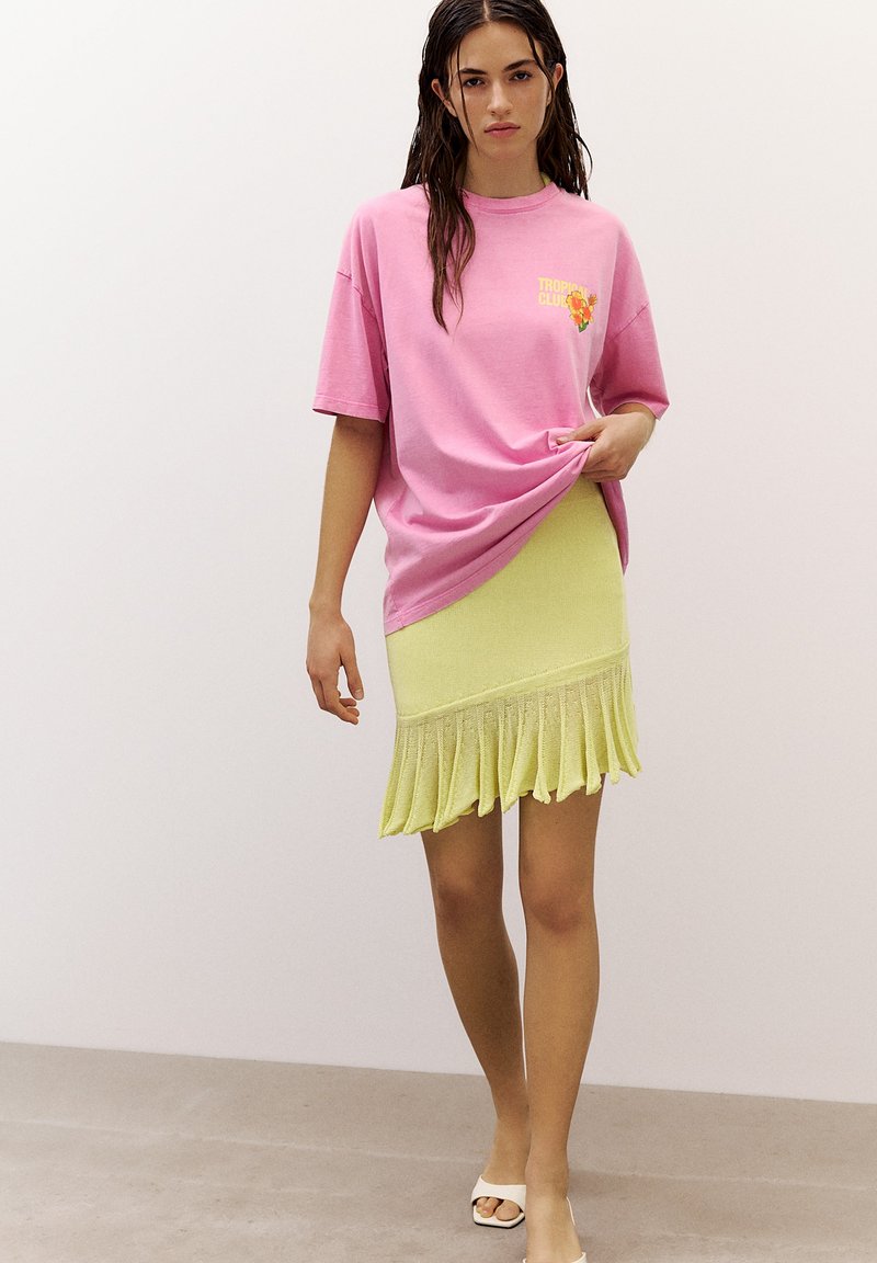 Woman with wet hair wears oversized pink T-shirt and pale yellow skirt with ruffled hem, standing against plain white background.