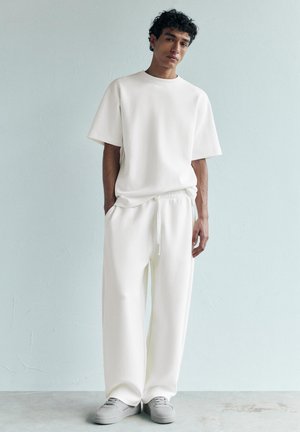 Junger Mann trägt ein lockeres weißes T-Shirt und weiße Hosen mit Kordelzug sowie graue Sneakers, steht vor einer hellgrauen Wand.