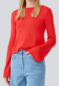 Haut rouge à manches longues avec un tricot texturé et un design de manches évasées, associé à un jean taille haute bleu clair, dévoilant un style décontracté.